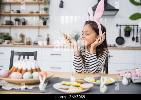 Lächelndes Kind mit Stirnband, das Osterkeks in der Nähe von Essen in der Küche ansieht Stockfoto