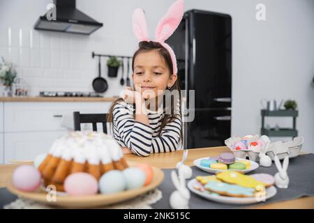 Ein Kind mit Hasenohren-Haarband, das Osterkuchen und Eier in der Küche ansieht Stockfoto