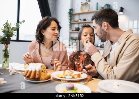 Mann füttert Tochter mit Osterkuchen in der Nähe ihrer Frau zu Hause Stockfoto