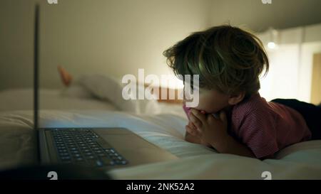 Das Kind schaute sich die Unterhaltungsmedien auf dem Laptop-Bildschirm an, der nachts im Bett lag. Ein hypnotisiertes Kind starrt auf einen blau leuchtenden Bildschirm Stockfoto