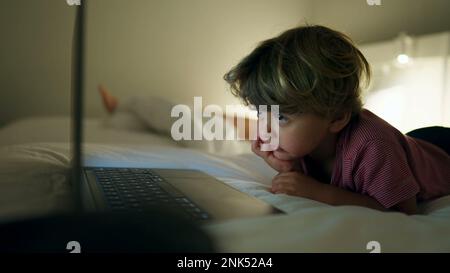 Das Kind schaute sich die Unterhaltungsmedien auf dem Laptop-Bildschirm an, der nachts im Bett lag. Ein hypnotisiertes Kind starrt auf einen blau leuchtenden Bildschirm Stockfoto