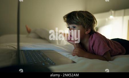 Das Kind schaute sich die Unterhaltungsmedien auf dem Laptop-Bildschirm an, der nachts im Bett lag. Ein hypnotisiertes Kind starrt auf einen blau leuchtenden Bildschirm Stockfoto