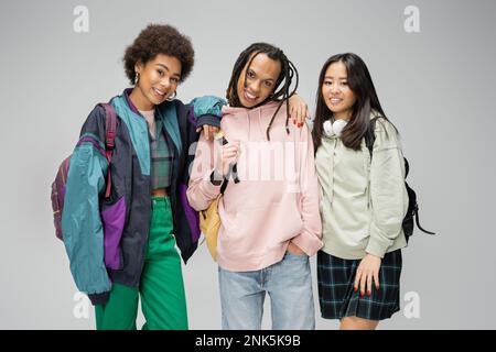 Hübsche multikulturelle Frauen, die sich neben einem trendigen Mann mit Dreadlocks und Rucksack auf grauem Stockbild postieren Stockfoto