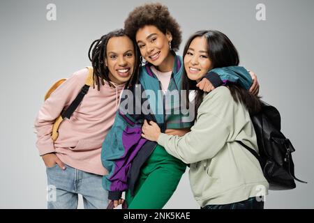 Fröhliche multikulturelle Freunde mit Rucksäcken, trendigen Kleidern und einem Lächeln vor der Kamera isoliert auf grauem Stockbild Stockfoto