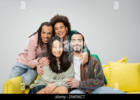 Zufriedene multiethnische Freunde, die trendige Kleidung tragen und neben der gelben Couch posieren, isoliert auf grauem Stockbild Stockfoto