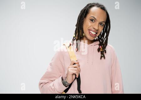 Ein fröhlicher, multiethnischer Mann mit Dreadlocks und Rucksack, der einen rosafarbenen Hoodie trägt und isoliert auf einem grauen Stockbild auf die Kamera schaut Stockfoto