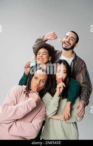 Aufregende multikulturelle Freunde in trendigen Freizeitkleidung, die Lutscher essen, isoliert auf grauem Stockbild Stockfoto