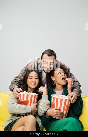 Ein fröhlicher bärtiger Mann, der Spaß hat, während er Popcorn in der Nähe von Frauen mit gemischten Rassen erreicht, die sich einen Film isoliert auf einem grauen Stockbild ansehen Stockfoto