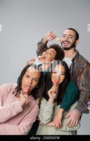 Fröhliche, multiethnische Freunde in trendigen Kleidern, die süße Lutscher essen, isoliert auf grauem Stockbild Stockfoto