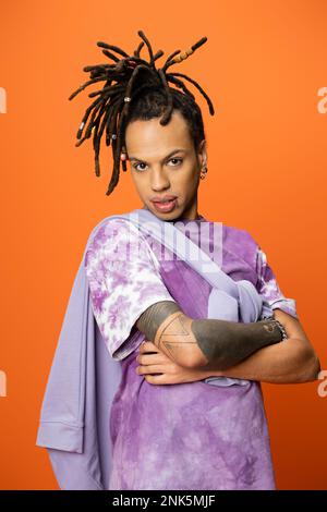 Trendiger, multiethnischer Mann mit Dreadlocks, der mit gekreuzten Armen auf orangefarbenem Stockbild posiert Stockfoto