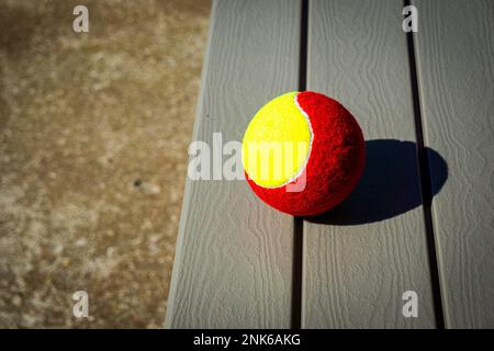 Ein rot-gelber Tennisball auf einer Holzbank. Sommeraktivität Stockfoto