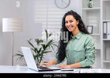 Porträt einer erfolgreichen, schönen Geschäftsfrau, einer hispanischen Frau im Heimbüro, die lächelt und in die Kamera schaut, einer Frau bei der Arbeit, die mit einem Laptop über die Tastatur tippt. Stockfoto