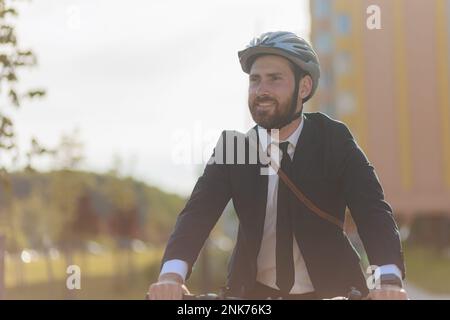 Glücklicher, bärtiger Geschäftsmann in Schutzhelm auf dem Weg zum Arbeitsplatz. Porträt eines jungen, fröhlichen Mannes in Anzug und Krawatte, der sich umsieht, während er reitet Stockfoto