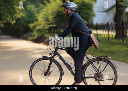 Konzentrierter männlicher Angestellter im Schutzhelm beim Fahrradfahren im Stadtpark. Seitenansicht eines bärtigen Managers mit Lederaktentasche, auf dem Weg zur Arbeit Stockfoto