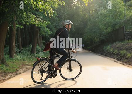 Eleganter Angestellter, der Fahrrad im Anzug fährt und am Park entlang nach Hause zurückkehrt. Lächelnder bärtiger Mann mit Schutzhelm, der Fahrrad fährt, während er zur Arbeit geht Stockfoto