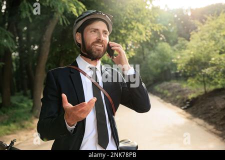 Freudiger bärtiger Mitarbeiter, der telefoniert, während er mit dem Fahrrad zur Arbeit fährt. Portrait eines glücklichen brunetten Geschäftsmannes im Schutzhelm-Gesten, während er macht Stockfoto