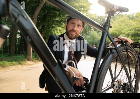 Ein hübscher bärtiger Mann in schwarzem Anzug hockt, weil er das Rad im Stadtpark überprüft hat. Portrait eines stilvollen männlichen Unternehmertums, der Fahrrad untersucht, während er schaut Stockfoto