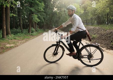 Lächelnder männlicher Angestellter in Schutzhelm auf dem Fahrrad entlang der bewaldeten Parkgasse. Seitenansicht des Arbeiters mit Lederaktentasche, Pendelverkehr mit bi Stockfoto