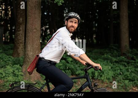 Ein freudiger Unternehmer auf dem Fahrrad, der am frühen Morgen durch den Stadtpark reitet. Seitenansicht eines braunen männlichen Arbeiters mit brauner Aktentasche, während er die Natur genießt Stockfoto