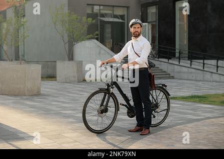 Junger Geschäftsmann pendelt mit dem Fahrrad nach Hause von der Arbeit in der Stadt. Seitlicher Blick auf den weißbärtigen Manager in Hemd und Helm, der mit modernem Stand steht Stockfoto