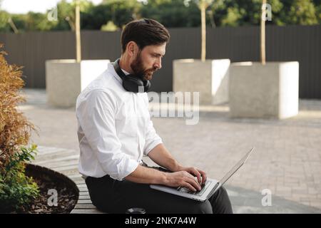 Konzentrierter bärtiger Mann tippt, während er ein Notebook zum Editieren von Dokumenten verwendet. Fokussierter Unternehmer mit Kopfhörern am Hals, der hart arbeitet, während er sitzt Stockfoto