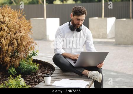 Ein seriöser, bärtiger Geschäftsmann, der auf einer Holzbank sitzt, mit einem Laptop auf den Knien. Ein weißer Mann mit Kopfhörern am Hals, der am Computer tippt und hart arbeitet Stockfoto