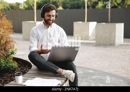Weißer Freiberufler im Bereich kabelloser Kopfhörer, der auf einem tragbaren Laptop sitzt, während er in der Stadt sitzt. Glücklicher Mann, der über das neue Projekt und die gesturi spricht Stockfoto