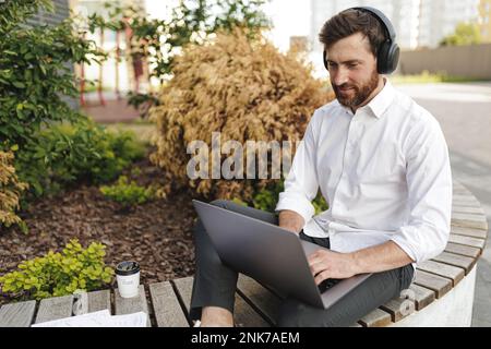 Gutaussehender, bärtiger Geschäftsmann in formeller Kleidung, der auf einem tragbaren Laptop tippt. Positiver Mann in Kopfhörern schaut aufmerksam auf den Bildschirm, während er sitzt Stockfoto