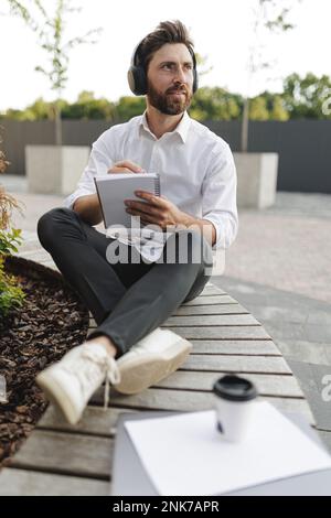 Eleganter Geschäftsmann im Bereich kabelloser Kopfhörer, der seine eigenen Ideen in Notizblock aufschreibt. Ein ernster Mann, der auf einer Bank sitzt und über neue Gedanken denkt Stockfoto