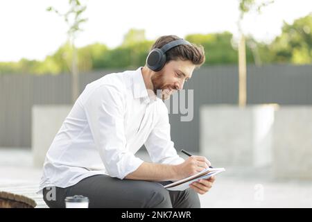 Seitenansicht eines weissen Büromitarbeiters mit Kopfhörern, der im Freien sitzt und kreative Ideen in ein Notizbuch schreibt. Dunkelhaariger Mann, der Musik hört und arbeitet Stockfoto