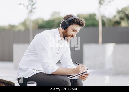 Selbstbewusster, gutaussehender Geschäftsmann mit Borsten, der im Stadtgebiet ruht und neue Ideen in Notizblock schreibt. Elegantes Männchen in weißem Hemd, das auf der Bank sitzt, während li Stockfoto