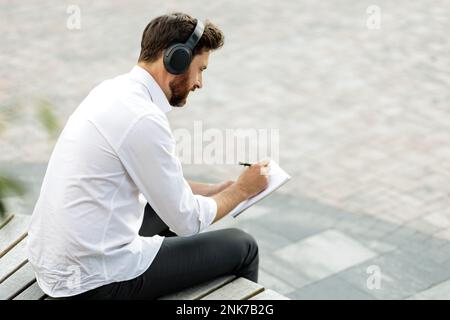 Eleganter, fokussierter Mann mit kabellosen Kopfhörern, der einen neuen Geschäftsplan in Notizblock schreibt. Dunkelhaarige Managerin mit Borsten, die Musik hört, während sie auf Woo sitzt Stockfoto