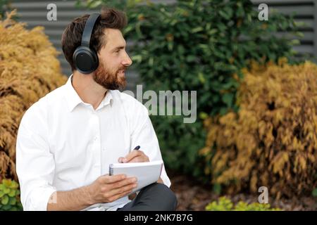 Seitenansicht eines gutaussehenden Mannes mit Borsten, der draußen sitzt und Tagebuch schreibt. Dunkelhaariger Freiberufler in Kopfhörern, der Musik hört, während er über sich nachdenkt Stockfoto