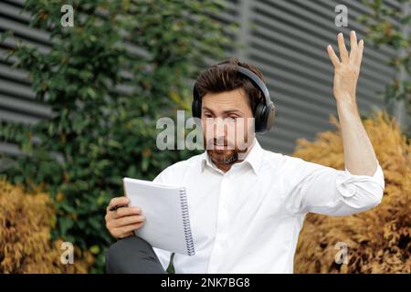 Emotionaler Mann mittleren Alters, der Hand hebt, während er draußen auf ein Notizbuch schaut. Portrait von aufgeregtem Brünetten in weißem Hemd und Kopfhörern, Gesten, Stockfoto