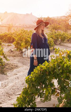 Wunderschöne blonde junge Frau mit blauem Rock und braunem Hut auf dem Friedhof - eine Plantage von Weinreben. Heller, warmer Effekt bei Sonnenaufgang im Frühling. Stockfoto