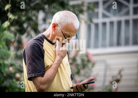 Der alte Mann schaut aufmerksam auf den Telefonbildschirm, steht auf der Straße neben einem Landhaus. Er hält sein Kinn mit einer Hand, in einem nachdenklichen Zustand. Verwenden von te Stockfoto