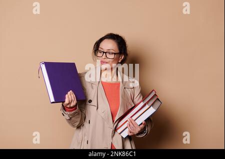 Schöne Frau mittleren Alters, Lehrerin in Brillenpädagogin, schaut sich Bücher auf beigefarbenem Hintergrund an Stockfoto