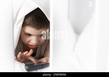 Ein 5-jähriger Junge spielt Spiele auf seinem Handy, während er im Bett liegt. Geräte im Bett vor dem Schlafengehen bei einem Kind Stockfoto