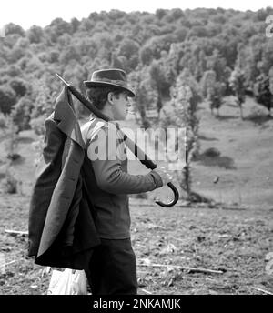 Kreis Suceava, Sozialistische Republik Rumänien, ca. 1980. Ein Mann, der mit einer Tasche, einem Regenschirm und einem Mantel auf dem Land herumläuft, während er eine Zigarette raucht. Stockfoto