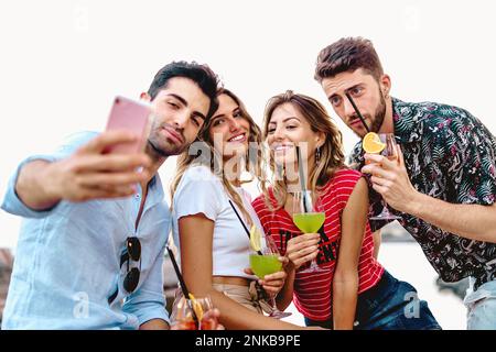 Vier junge Leute, zwei Männer und zwei Frauen, die sich zur Happy Hour draußen amüsieren, Cocktails trinken und Selfies machen, während sie lächeln und tog halten Stockfoto