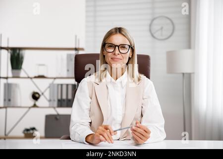 Lächelnde Geschäftsfrau mittleren Alters, die sich Webcam-Videokonferenzen ansieht, in einem virtuellen Chat-Meeting anruft und von dort aus einen Webinar-Kurs abhält Stockfoto