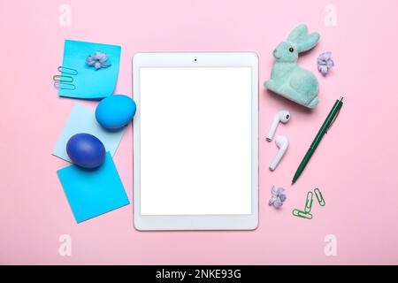 Tablet-Computer mit Büromaterial, Ostereiern, Kaninchen und Blumen auf pinkfarbenem Hintergrund Stockfoto