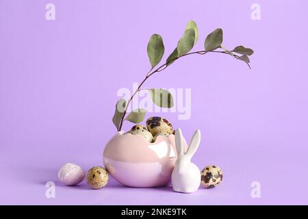 Dekorativer Korb mit Osterwachteleiern, Häschen- und Eukalyptuszweig auf lila Hintergrund Stockfoto