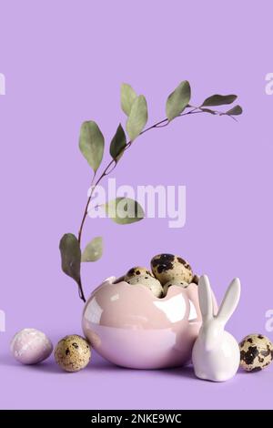 Dekorativer Korb mit Osterwachteleiern, Häschen- und Eukalyptuszweig auf lila Hintergrund Stockfoto