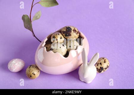 Dekorativer Korb mit Osterwachteleiern, Häschen- und Eukalyptuszweig auf lila Hintergrund Stockfoto