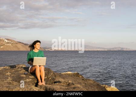 Lächelnde Frau mit Laptop, die auf Felsen am Meer sitzt Stockfoto