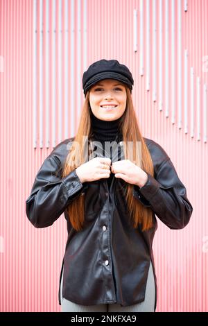 Glückliche Rothaarige, die Mütze und Jacke vor der rosa Wand trägt Stockfoto