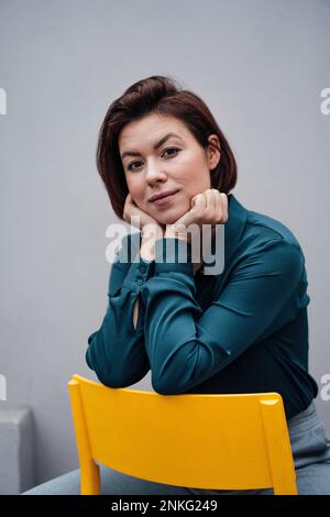 Selbstbewusste junge Geschäftsfrau, die auf einem Stuhl vor der Wand sitzt Stockfoto