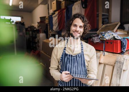 Glücklicher Besitzer trägt eine Schürze im Stehen mit einem Tablet-PC im Lagerraum Stockfoto