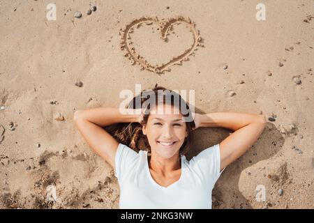 Lächelnde Frau mit Händen hinter dem Kopf, die am Strand auf Sand liegt Stockfoto